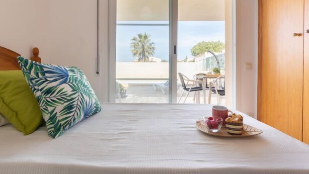 Un dormitorio con cama, almohada verde, cubrecama blanco y bandeja de desayuno con pastel, taza y bayas.