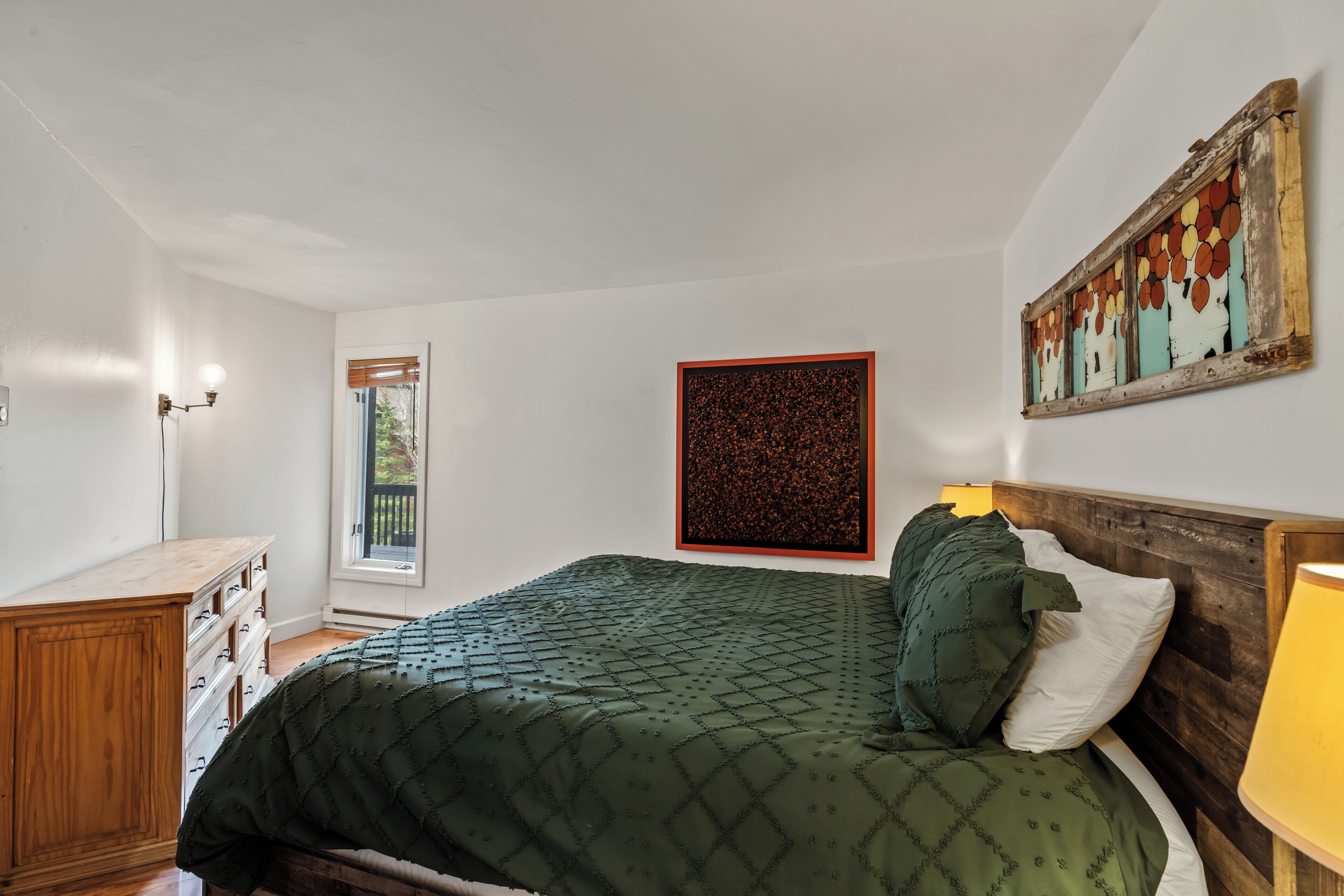 A bedroom with a bed, a wooden dresser, a window, and a wall-mounted artwork.