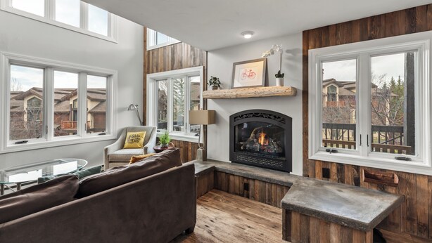 Una sala de estar acogedora con chimenea, detalles de madera, un sofá y ventanas amplias con vistas al vecindario.