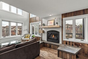 A cozy living room with a fireplace, wooden accents, a sofa, and large windows offering a view of the neighborhood. - Blocks to the Slopes | Fireplace & Winter Shuttle (Steamboat Springs)