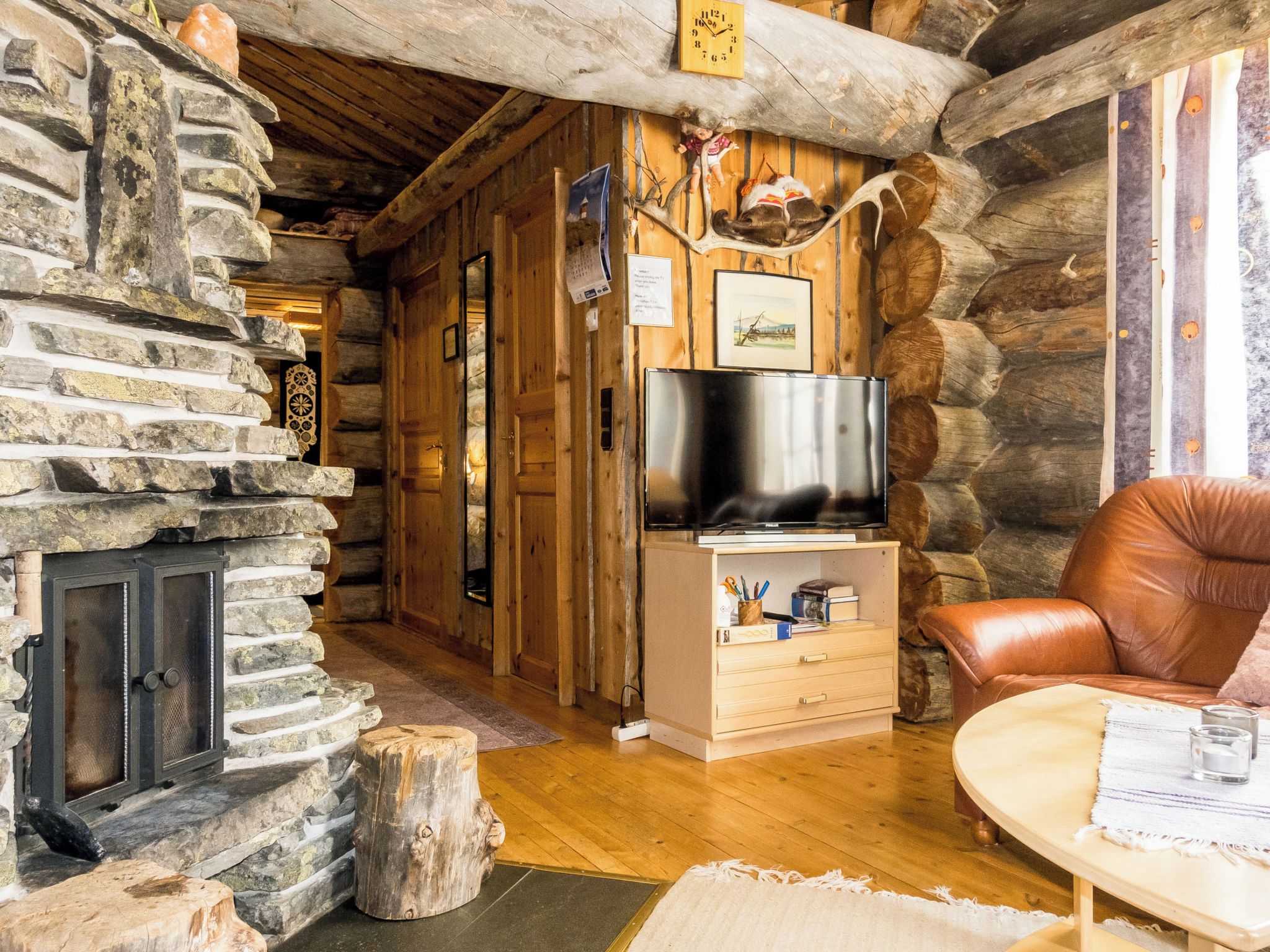 A log cabin interior with a stone fireplace, wooden walls, a television on a wooden cabinet, and a leather chair.