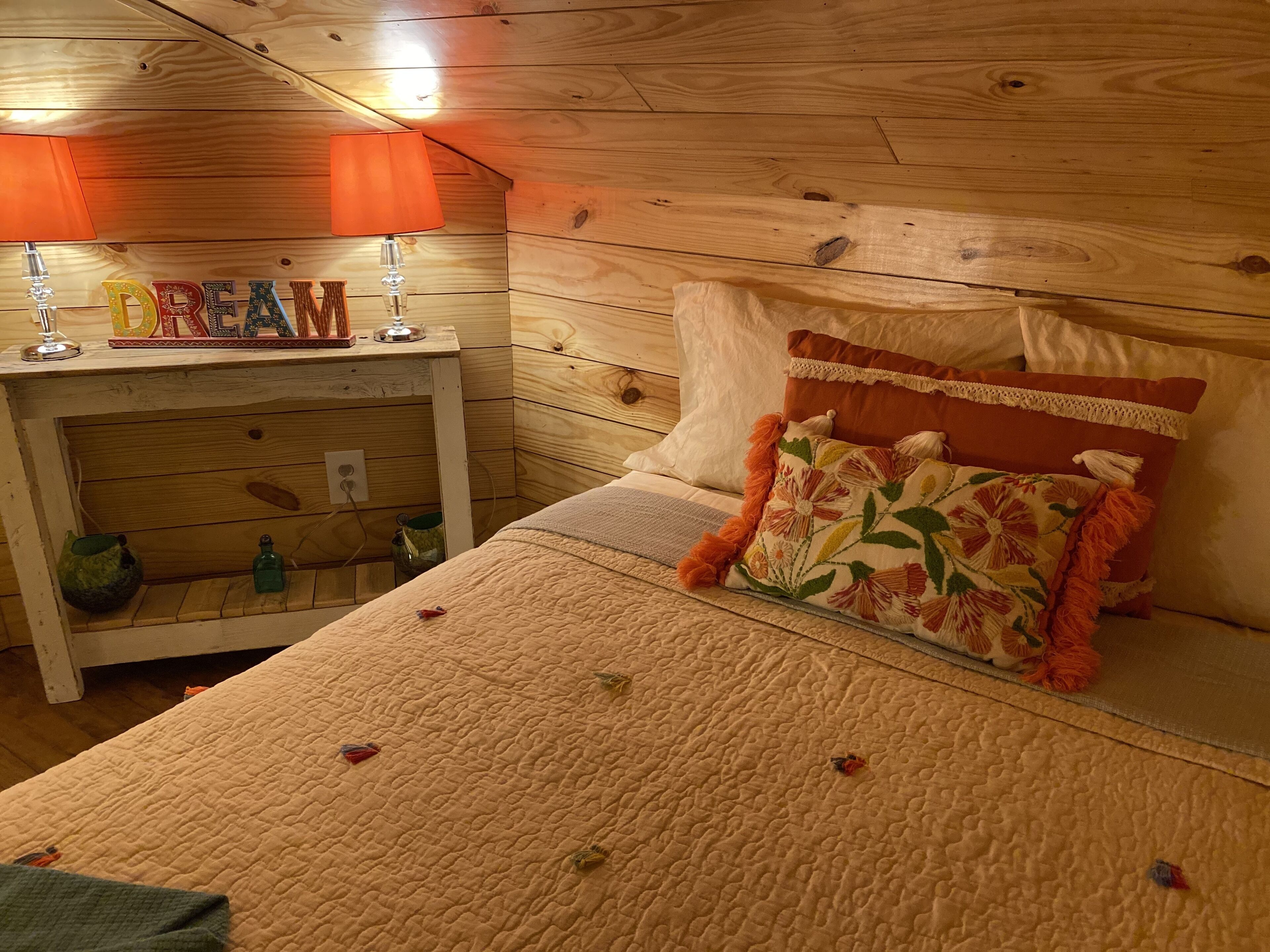 A cozy bedroom with a wooden ceiling, a nightstand with a lamp, a bed with floral pillows, and a 'DREAM' sign.