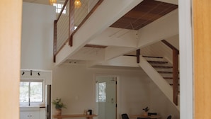 Een ruime woonkamer met een grote hoekbank, een houten salontafel en een eettafel met stoelen. Een trap met houten leuningen leidt naar een hoger gelegen verdieping.