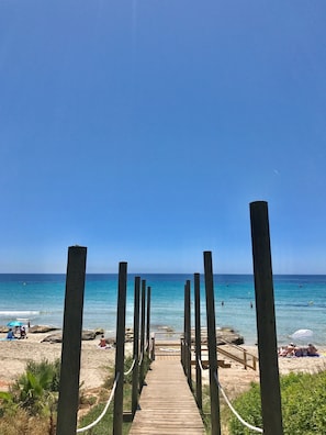 Un sendero de madera que lleva a una playa con agua azul cristalina y algunas personas a lo lejos.