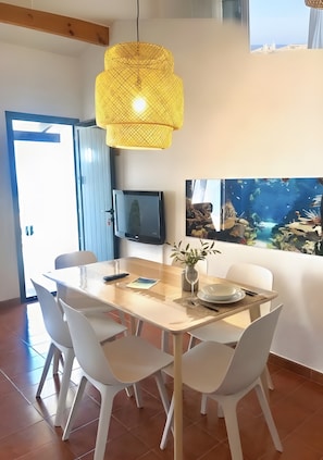 Un comedor con una mesa de madera, sillas blancas, una lámpara colgante amarilla, un televisor y un acuario en la pared.