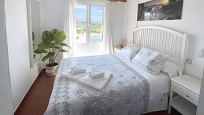 Une chambre à coucher avec un lit, une table de chevet, une plante en pot, un ventilateur de plafond et une fenêtre avec des rideaux.