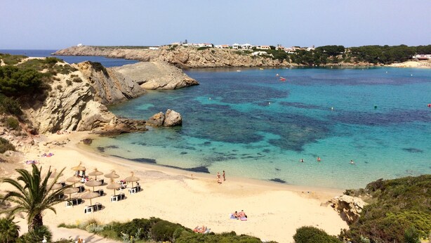 Une plage côtière aux eaux cristallines, parsemée de rochers et bordée d’une étendue de sable où sont installés des parasols et des chaises longues.