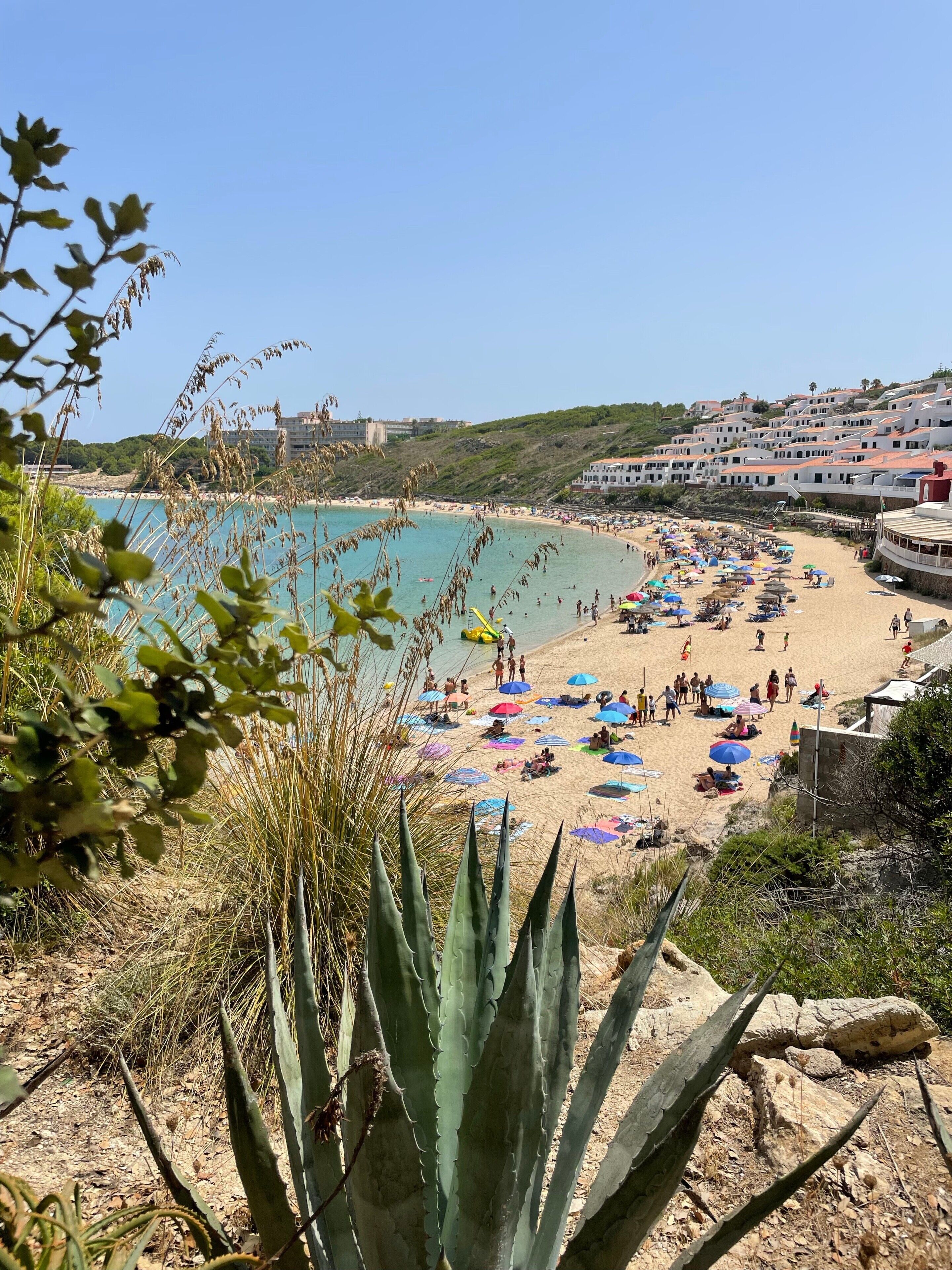 Escena de playa costera con gente, sombrillas y edificios al fondo.