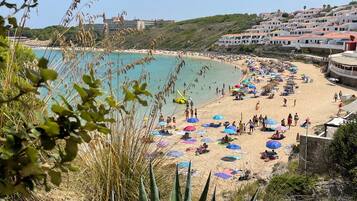 Escena de playa costera con gente, sombrillas y edificios al fondo.