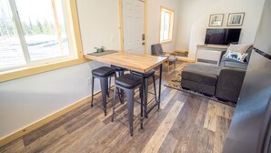 A compact living space with a wooden dining table, two bar stools, a gray sofa, and a television.