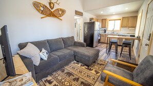 A modern living room with a gray sectional sofa, a wooden coffee table, and a patterned rug. A television and books are on the table. The kitchen in the background has wooden cabinets and a stainless steel refrigerator.