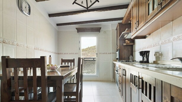 A kitchen with wooden cabinets, a dining table with chairs, a clock on the wall, and a view of the outdoors through a door. - Vacation Home "Apartamento Vistas de Icod" with Mountain View, Sea View & WiFi (Icod de los Vinos)