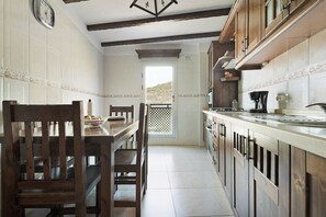 Une cuisine avec des meubles de rangement en bois, une table à manger avec des chaises, une horloge au mur et une vue sur l’extérieur par une porte.