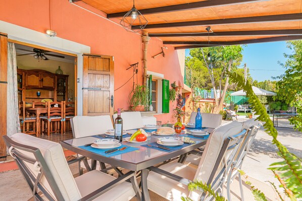Un área de comedor al aire libre con una mesa dispuesta para dos personas, rodeada de sillas, bajo una estructura sombreada.