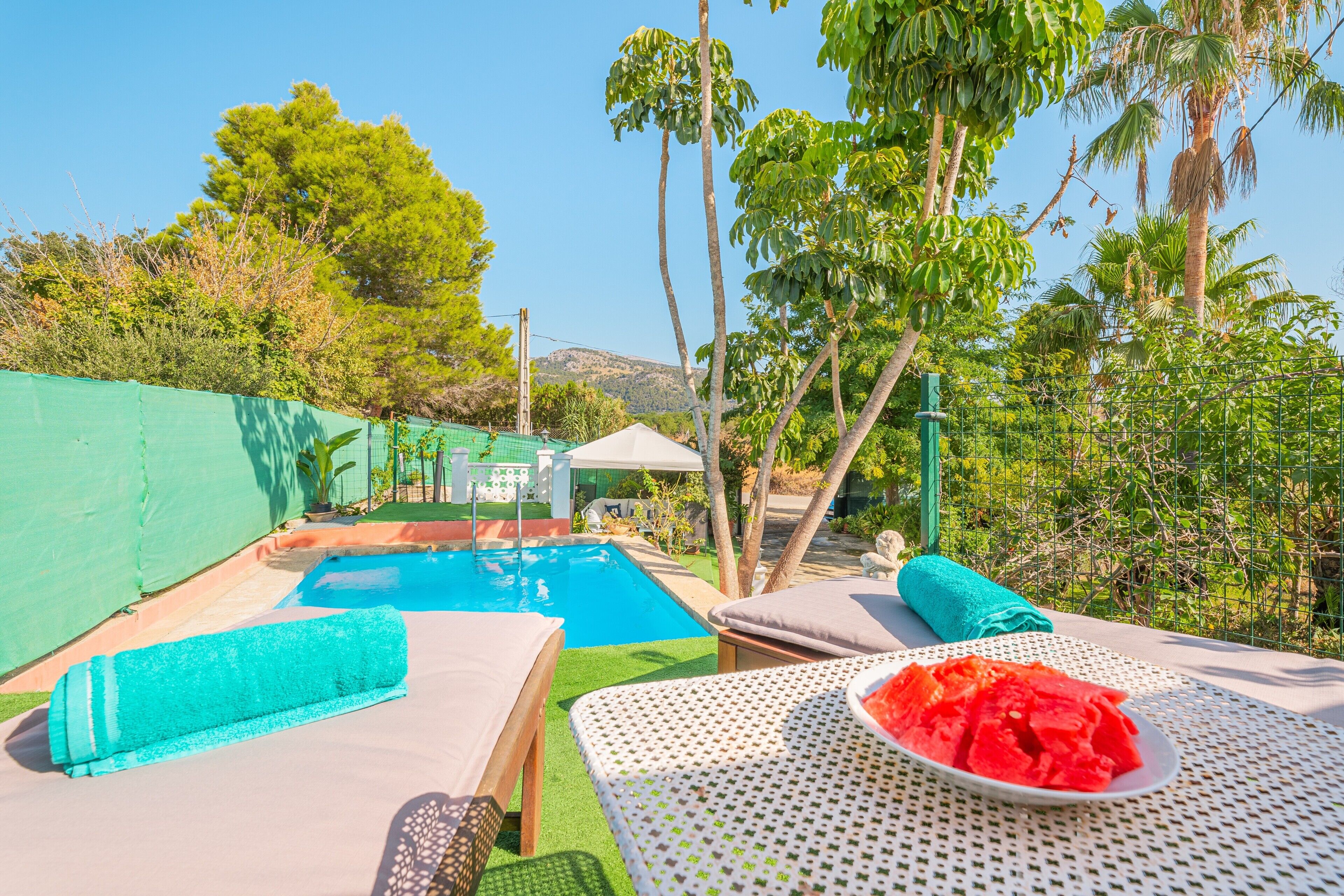 A poolside area with lounge chairs, a table with watermelon, and a view of a pool and greenery.