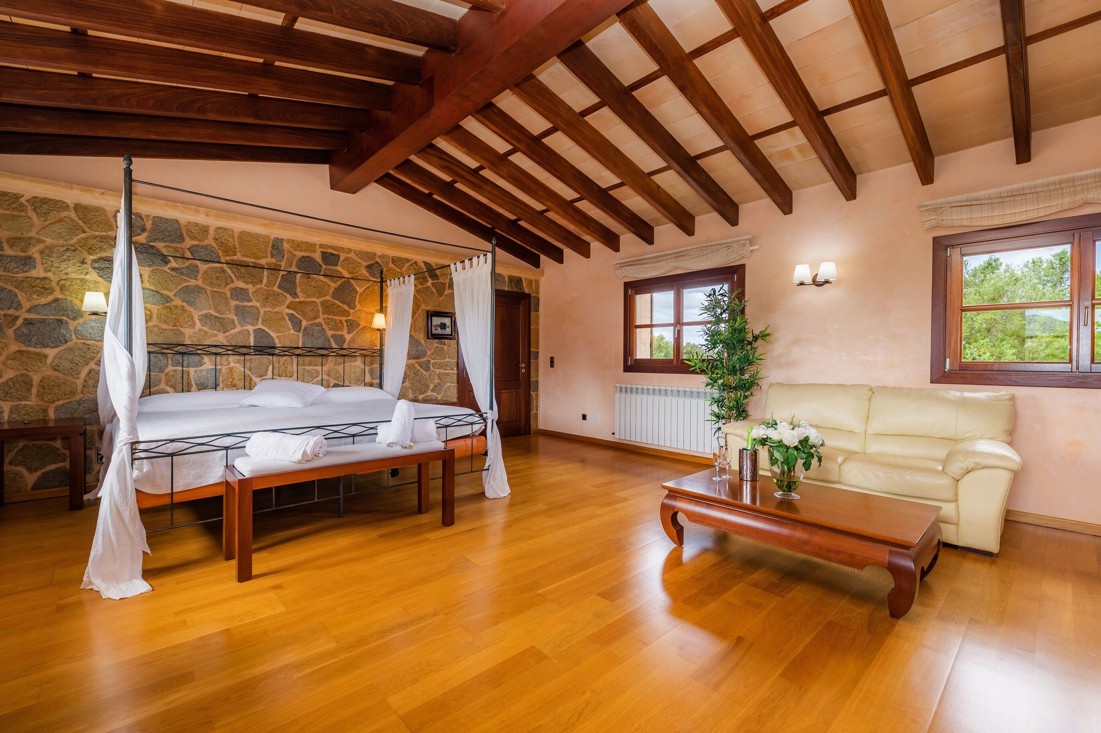 Una habitación con pared de piedra, vigas de madera en el techo, una cama con dosel, un banco, un sofá y una ventana con vistas.