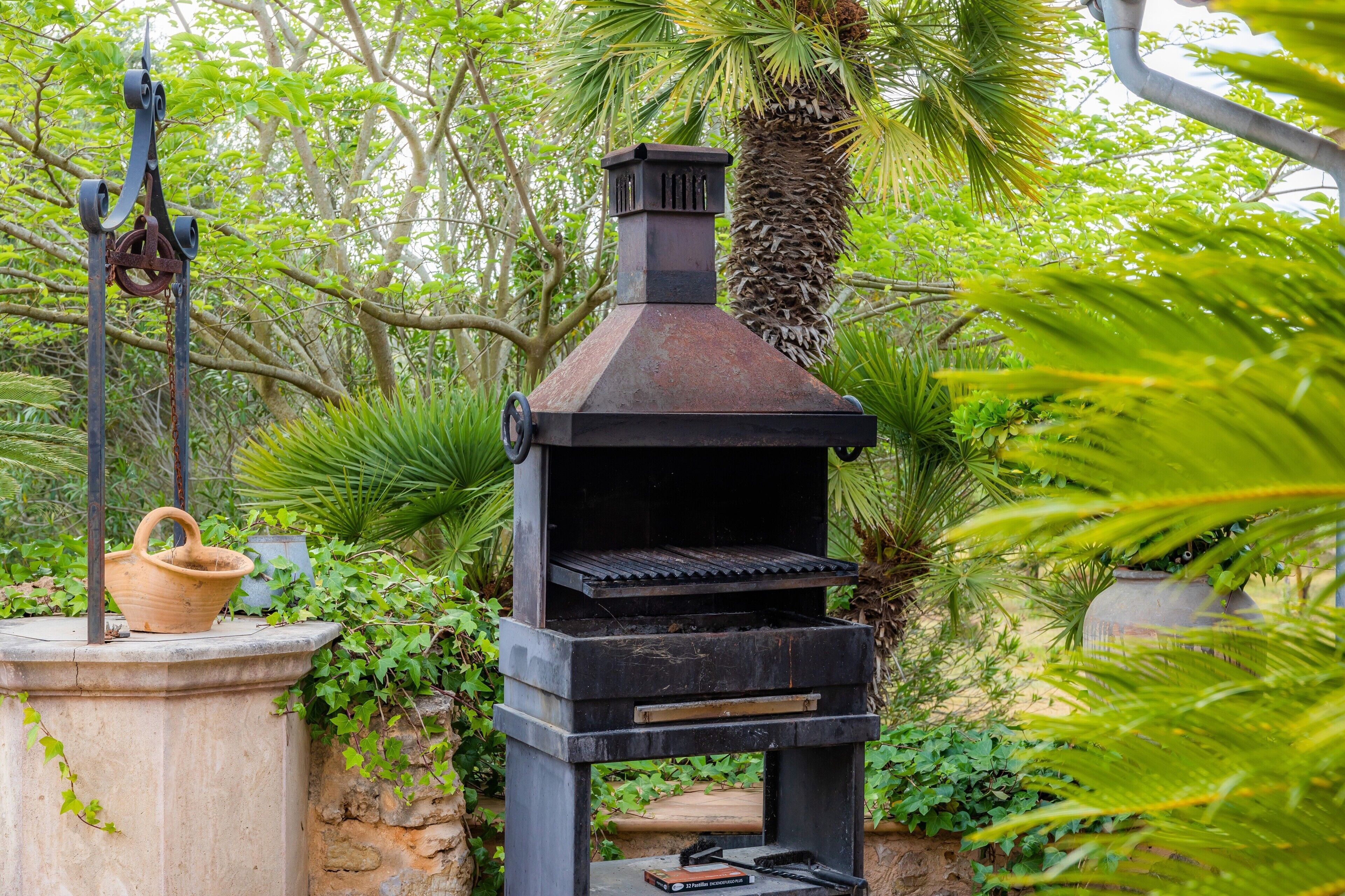 Una barbacoa negra per a l'exterior amb una xemeneia, envoltada de vegetació abundant i una base de pedra.