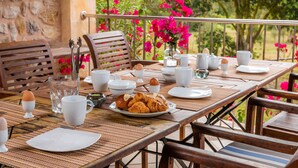 Mesa de café da manhã posta com ovos, croissants e xícaras de café, cercada por cadeiras e uma parede de pedra.