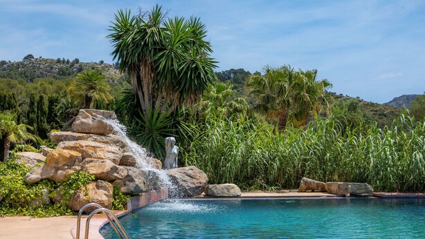 Piscina com cascata, rodeada por vegetação tropical e montanhas ao fundo.