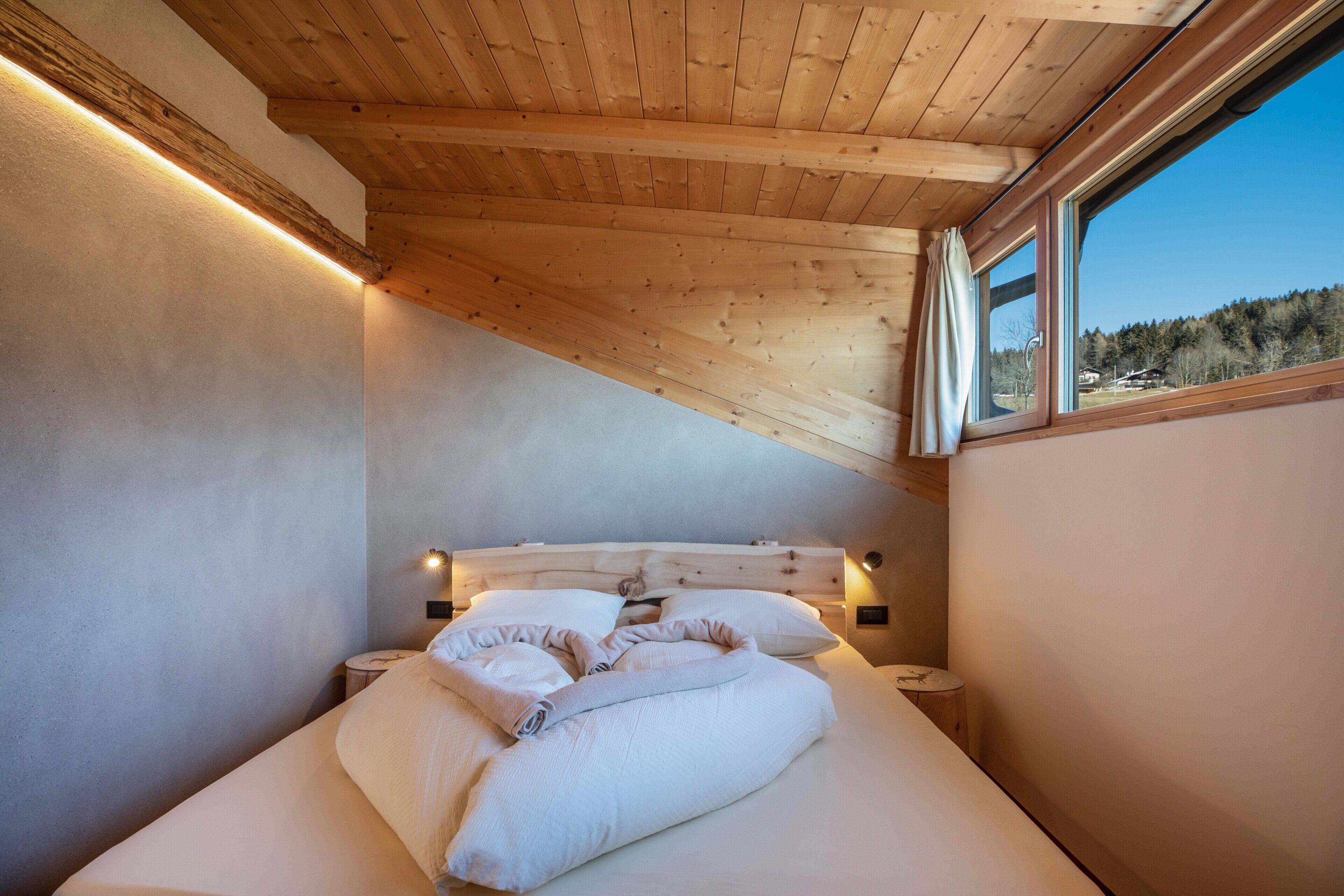 A bedroom with a wooden ceiling, a large bed with white bedding, bedside lamps, and a window with a view of a forest.