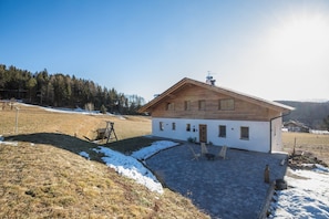 Ein modernes Haus mit Satteldach, einer gepflasterten Terrasse und Blick auf einen bewaldeten Bereich.