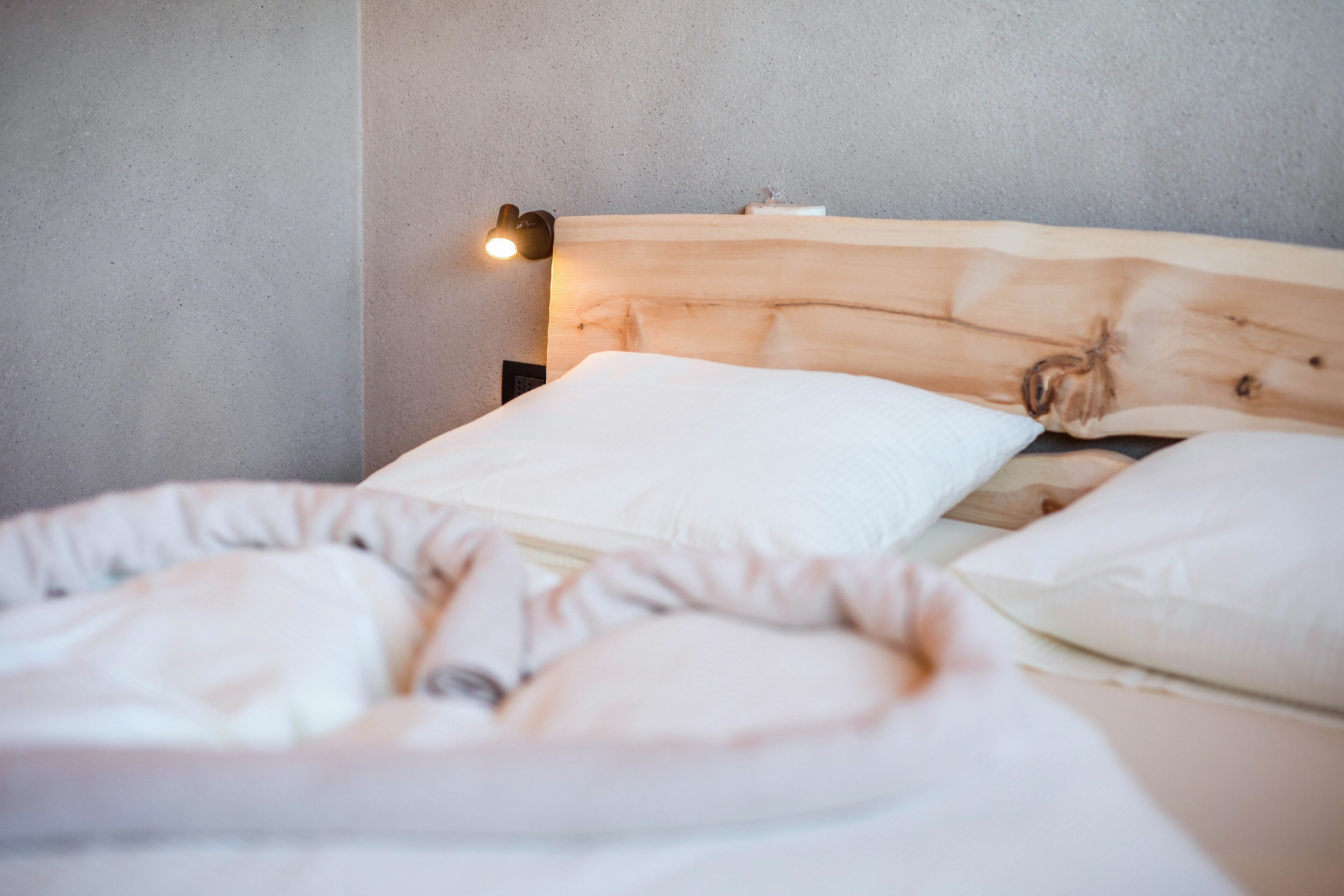 Una cama con cabecera de madera, almohadas blancas y una lámpara fijada a la pared.