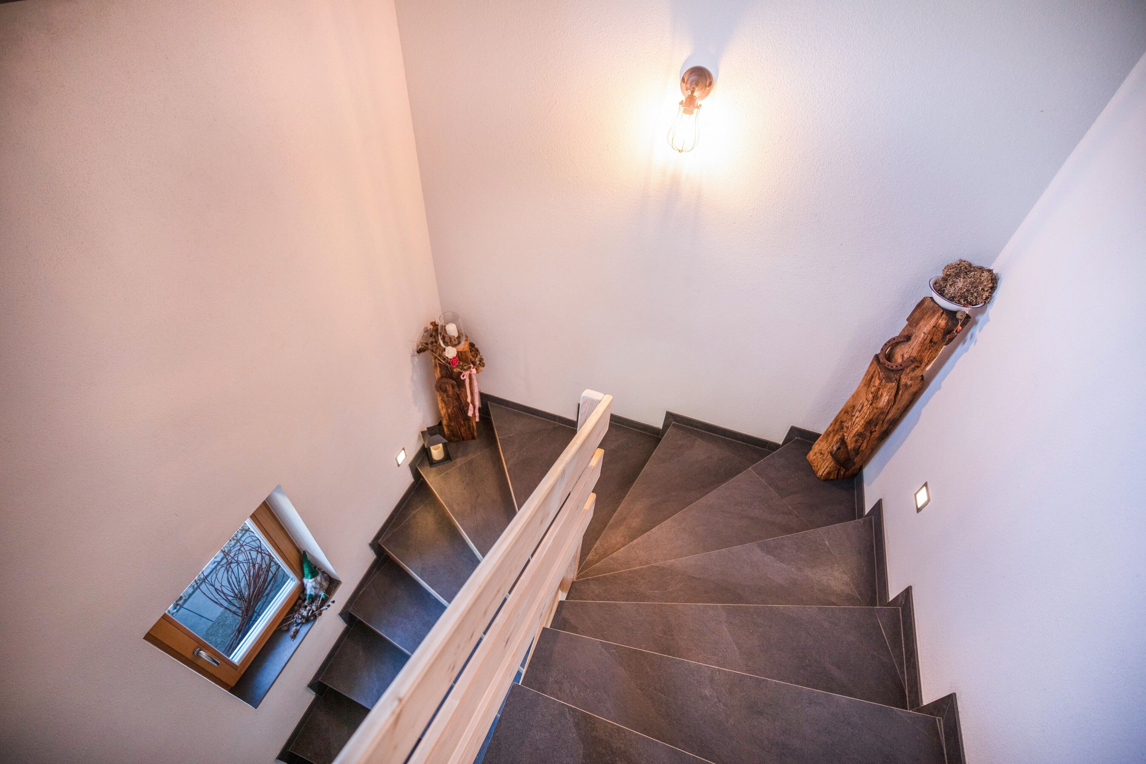Un escalier recouvert de carreaux sombres, une main courante en bois, un luminaire fixé au mur et une petite fenêtre donnant sur l’extérieur.