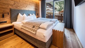 Ein modernes Hotelzimmer mit einem Holz-Kopfteil, einem großen Bett, einem Nachttisch aus Holz und Blick auf eine Holzterrasse und Berge.