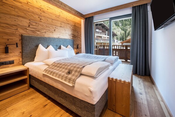 A modern hotel room with a wooden headboard, a large bed, a wooden nightstand, and a view of a wooden deck and mountains.