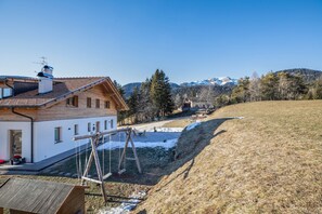 A wooden house with a sloped roof, a swing set, and a view of snow-capped mountains.