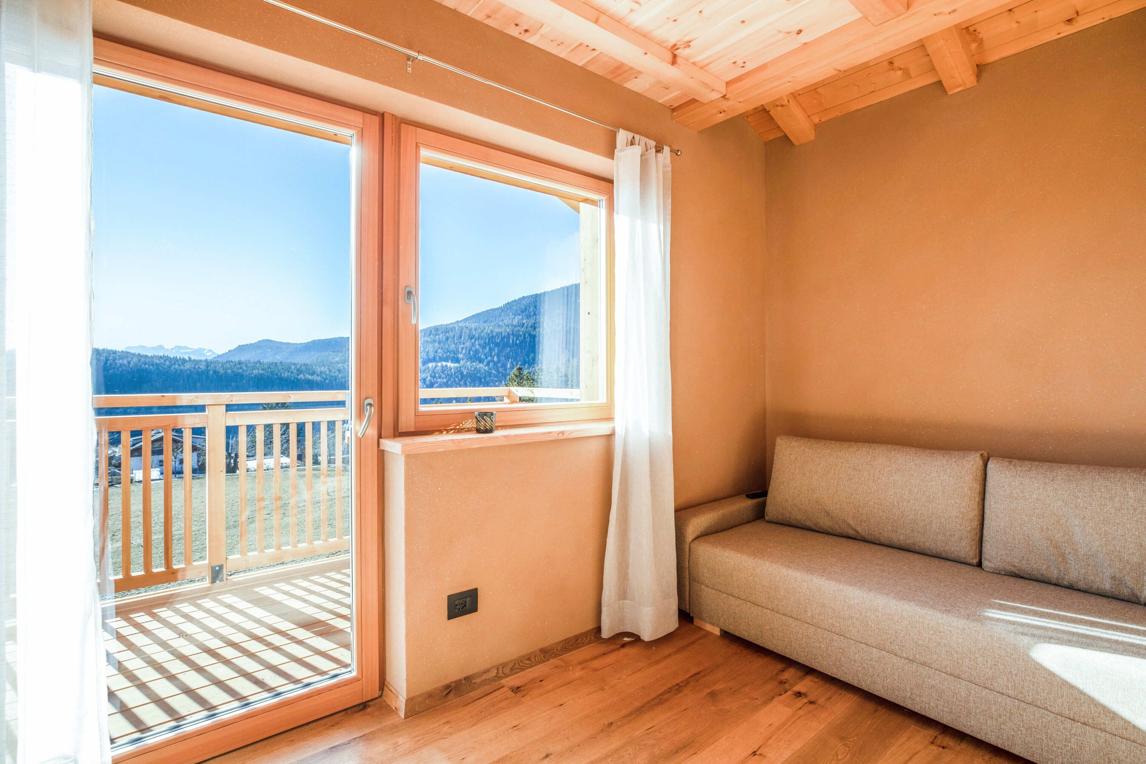 Quarto com piso de madeira, sofá, janela com vista para montanhas e varanda com grade de madeira.