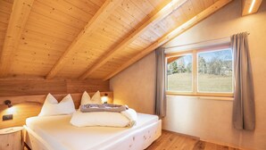 Ein Schlafzimmer einer Holzhütte mit Bett, Kissen, einem Nachttisch, einem Fenster mit Vorhängen und Blick auf eine Wiese.
