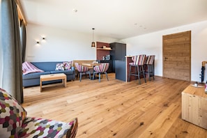 A modern living room with a sofa, a dining table with chairs, a wooden coffee table, and a kitchen area with cabinets and a hanging light.