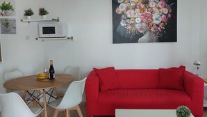 A living room with a red sofa, a white coffee table, a dining table with chairs, a microwave, and a painting of flowers. - Apartamento Sol y Playa with Pool, Wi-Fi, Terrace & Garden (Puerto del Carmen)