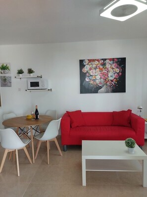 A living room with a red sofa, a white coffee table, a dining table with chairs, a microwave, and a painting of flowers.