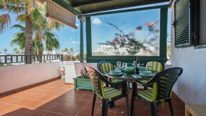 A covered outdoor dining area with a table set for four, surrounded by chairs with striped cushions, a view of palm trees, and a clear sky. - Apartamento Sol y Playa with Pool, Wi-Fi, Terrace & Garden (Puerto del Carmen)