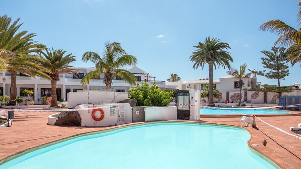 A swimming pool with a lifebuoy, palm trees, and a building with a white facade. - Apartamento Sol y Playa with Pool, Wi-Fi, Terrace & Garden (Puerto del Carmen)
