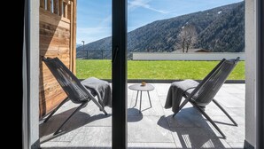 A room with a sliding glass door, two lounge chairs, a small round table, and a view of a mountainous landscape. - Apartment "Mulin Hilda" next to the Skiing Area with Mountain View, Sauna & Garden (Vals)