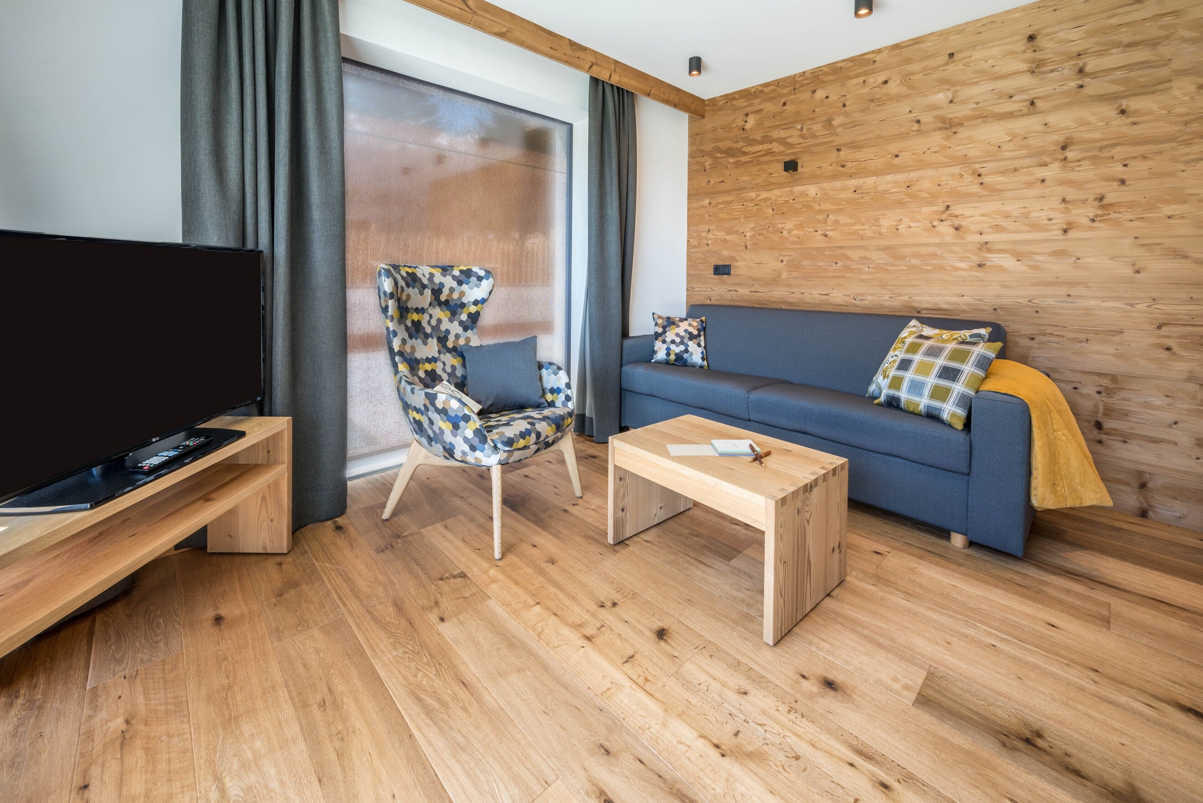 Una sala de estar moderna con un televisor de pantalla plana, un sillón estampado, un sofá azul y una mesa de centro de madera.