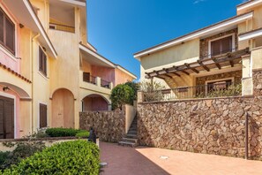 A multi-story residential building with balconies, a stone wall, and a paved courtyard. - Apartment 'Isabel' with Shared Pool, Wi-Fi and Air Conditioning (villasimius)