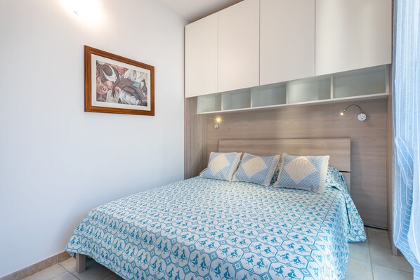 A bedroom with a bed, a headboard, a wall-mounted shelf, a framed picture, and a bedside lamp.