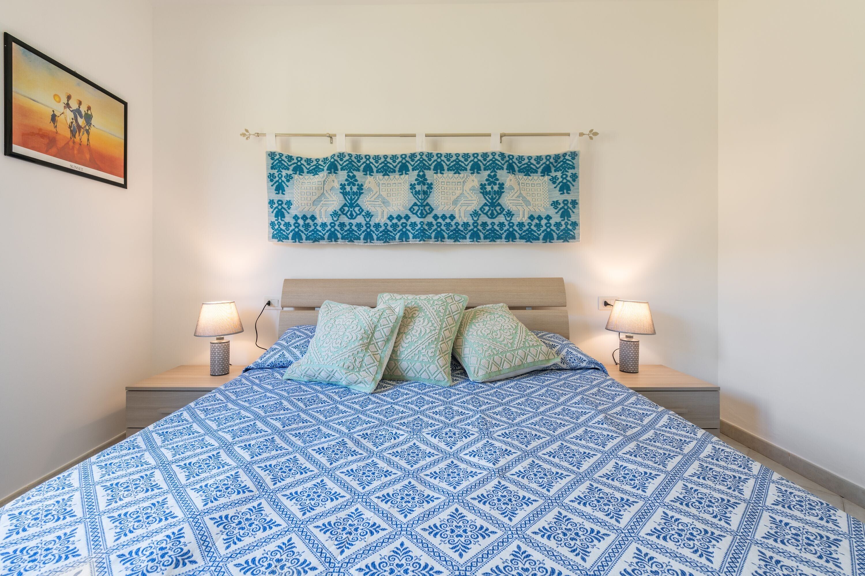A bedroom with a patterned bedspread, a headboard with a hanging textile, two bedside lamps, and a framed picture on the wall.