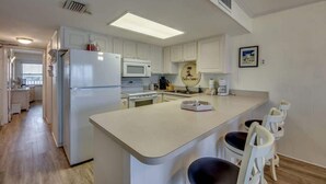 A kitchen with white cabinets, a refrigerator, a microwave, a coffee maker, and a small island with bar stools. - Gateway Villas #795 Cozy Beachfront Condo (Fort Myers Beach)