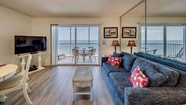 A living room with a sofa, a television, a dining table, and chairs. There is a balcony with a view of the sea. - Gateway Villas #795 Cozy Beachfront Condo (Fort Myers Beach)