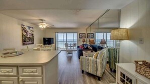 A modern living room with a sofa, a coffee table, and a TV. There is a kitchen area with a counter and cabinets. - Gateway Villas #795 Cozy Beachfront Condo (Fort Myers Beach)