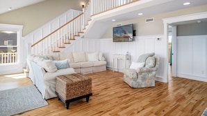 A spacious living room with a wooden floor, a beige sofa, a patterned armchair, and a wicker ottoman. A staircase with white railings leads to an upper level. - "It Was A Good Day" - 2nd Row Home, 4 Bedrooms / 4.5 Bathrooms (Holden Beach)