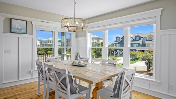 A dining room with a wooden table and white chairs, large windows with a view of houses, and a chandelier. - "It Was A Good Day" - 2nd Row Home, 4 Bedrooms / 4.5 Bathrooms (Holden Beach)