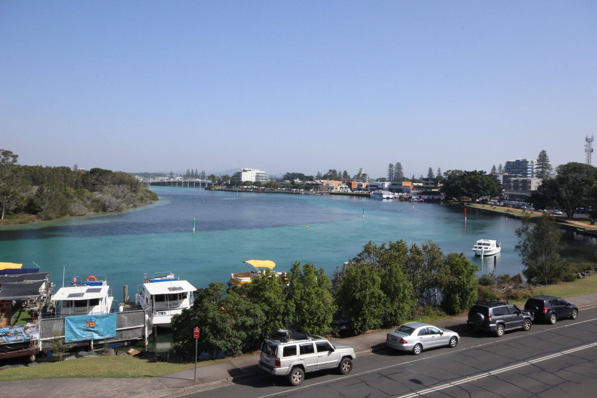 A marina with boats docked, a bridge in the background, and a road with parked cars.