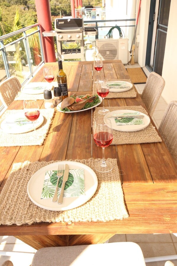 A wooden dining table set with plates, cutlery, and wine glasses, surrounded by wicker chairs. - Spinnaker Unit 201 (Forster)