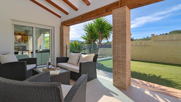 Un patio couvert aménagé avec des meubles en osier, une porte vitrée donnant sur la cuisine et une vue sur une pelouse verdoyante et des arbres.
